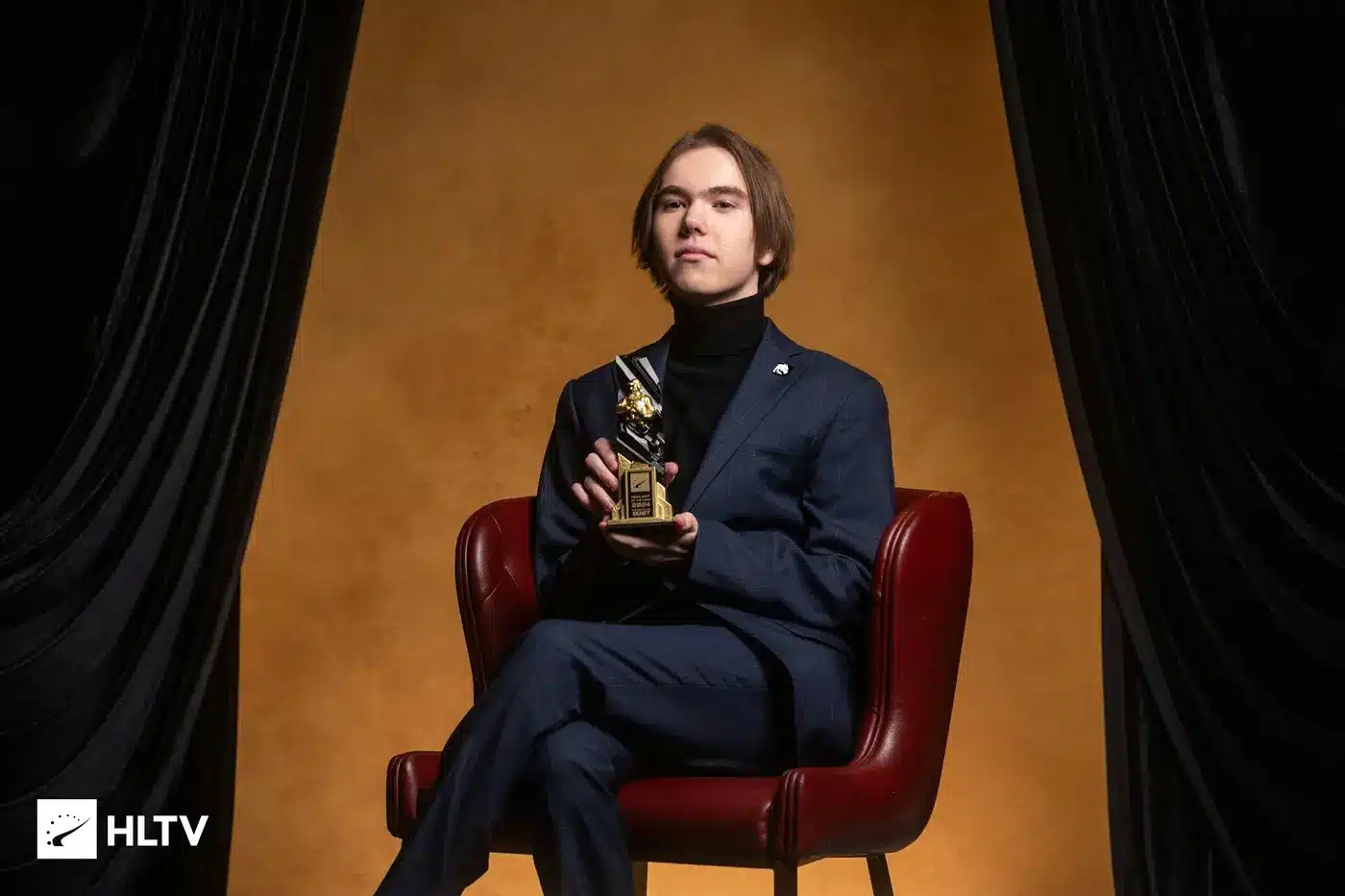 Danil Kryshkovets, known as donk, wearing a blue suit and holding an award while sitting in a red chair