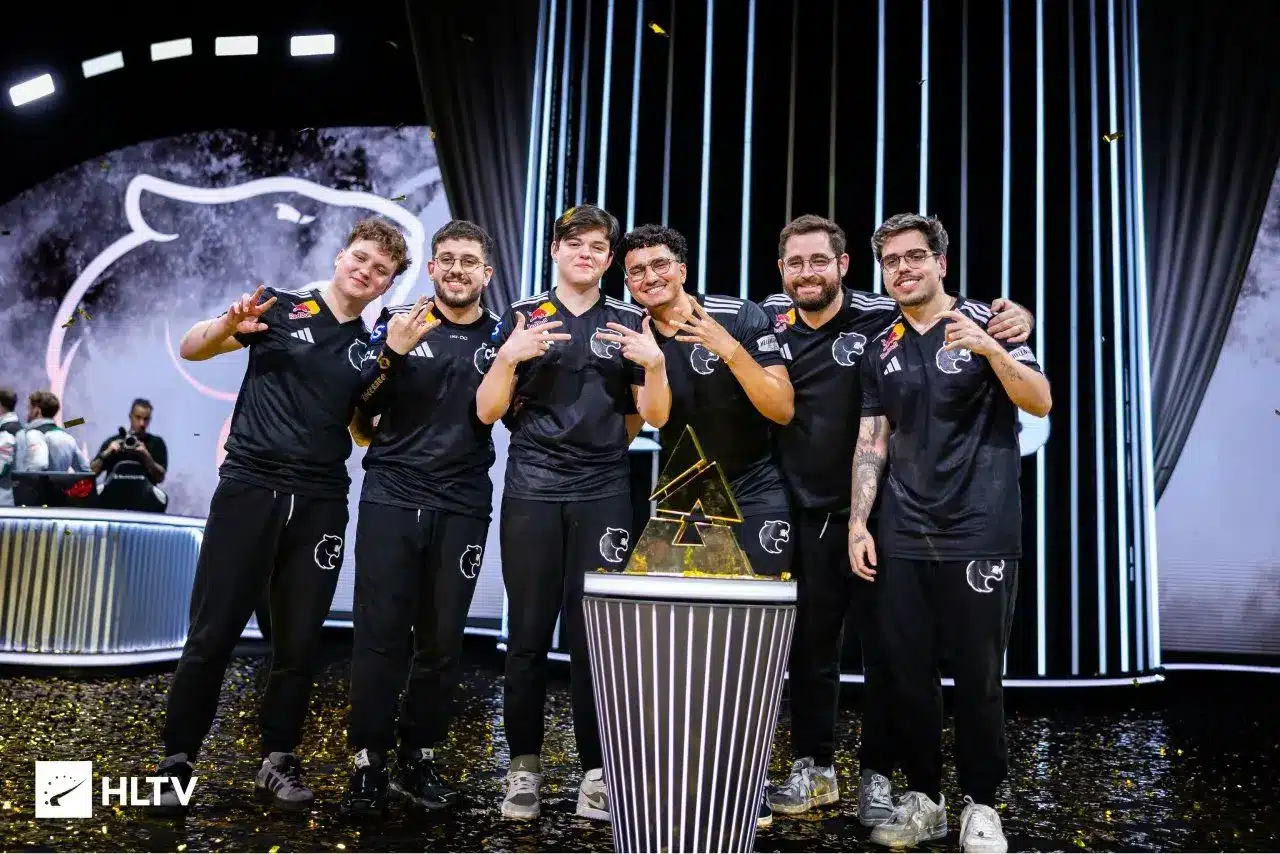 Six members of the FURIA esports team in black jerseys posing with a gold trophy on a stage with confetti.