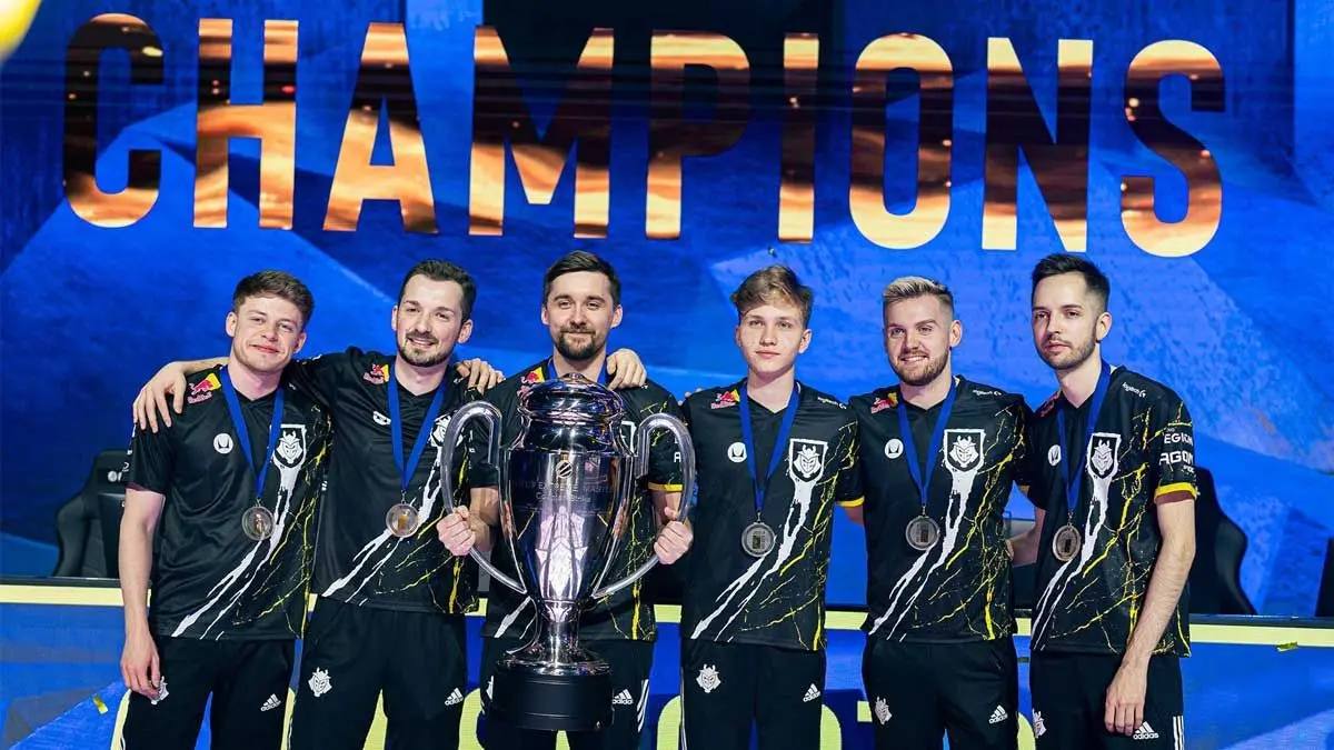G2 Esports Counter-Strike team standing together holding a silver trophy in front of a Champions backdrop